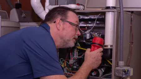 Technician with glasses inspecting and repairing HVAC system wiring using a red flashlight indoors.