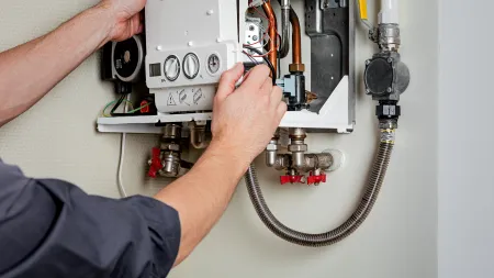 Technician repairing a wall-mounted boiler with visible pipes, dials, wires, and valves in a home.