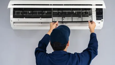 Technician in blue uniform cleaning the filter of a wall-mounted air conditioner indoors against a gray wall.