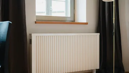 White vertical panel radiator under a window with dark curtains and a standing lamp in a cozy room corner
