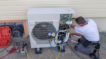 Technician repairing an HVAC unit outside a house using gauges and various tools on concrete ground.