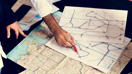 People analyzing and marking transportation route maps on a table with printed maps and documents around.