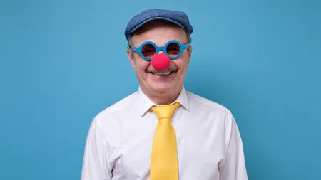 Smiling elderly man wearing blue cap, blue round glasses, red clown nose, white shirt, and yellow tie on blue background