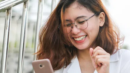 Smiling woman with glasses using smartphone outdoors near metal railing, showing excitement and joy.