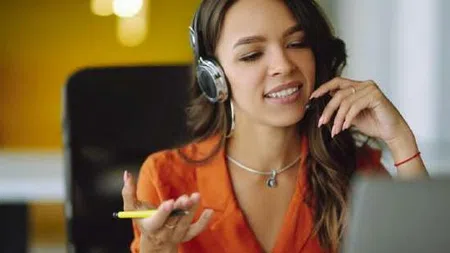 Young woman in orange shirt wearing headphones and holding a pen while looking at a laptop screen.