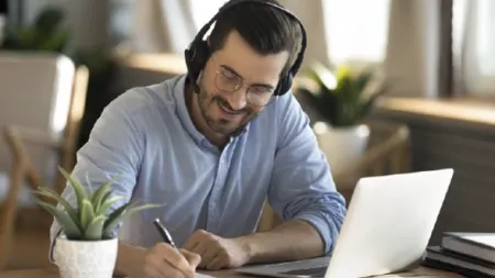 Young man with headset writing notes while working on laptop at a bright home office desk