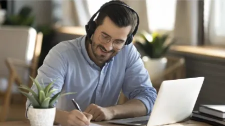 Smiling man wearing headphones writing notes with pen while looking at laptop in bright workspace.