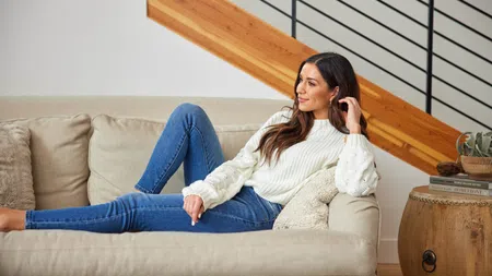 Woman lounging on beige couch in cozy white sweater and jeans in modern living room with wooden accents.