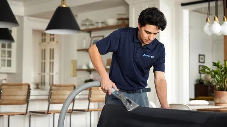 Cleaning professional in navy shirt using upholstery cleaner on black couch in modern home interior