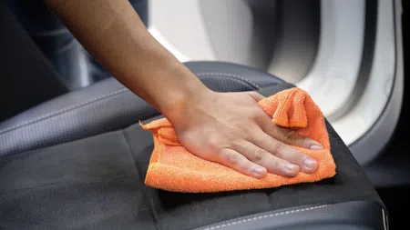 Person cleaning black car seat with orange microfiber cloth near car door interior.