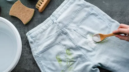 Hand spraying baking soda on green stain on white denim jeans for stain removal with cleaning tools nearby