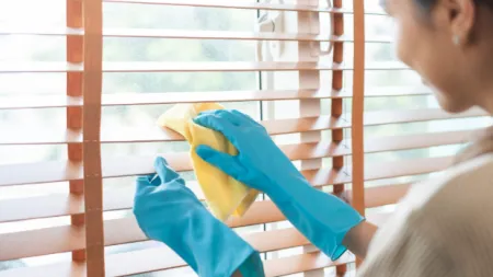 Person wearing blue gloves cleaning wooden window blinds with a yellow cloth in bright natural light.