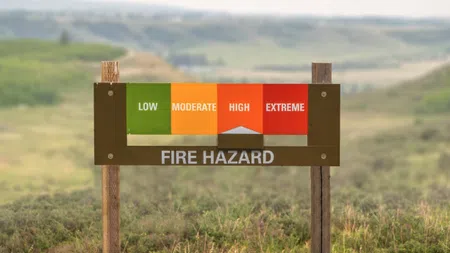 Outdoor fire hazard sign showing levels from low to extreme with arrow pointing at high risk in grassy landscape.