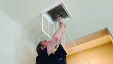 Technician installing or repairing wiring in a ceiling access panel inside a room with white walls and ceiling.