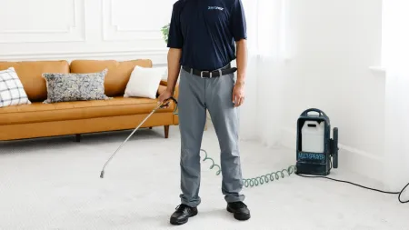 a male Zerorez technician holding a sprayer, applying Premium fiber protector to a light gray living room carpet