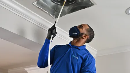 Technician in blue uniform and mask cleaning an air duct with a specialized tool indoors.