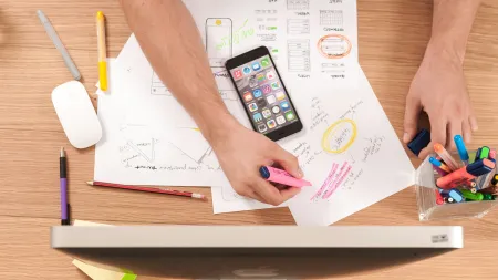 Person highlighting notes on printed charts with smartphone and computer on wooden desk workspace.