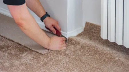 Hands cutting beige carpet along the wall corner with a utility knife during flooring installation.