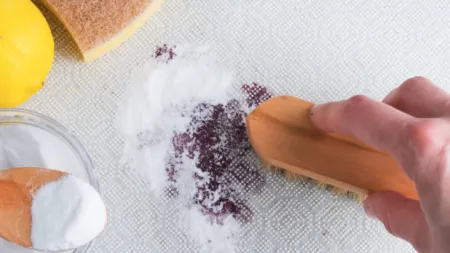 Hand scrubbing red wine stain on fabric using baking soda, brush, lemon, and sponge for stain removal.