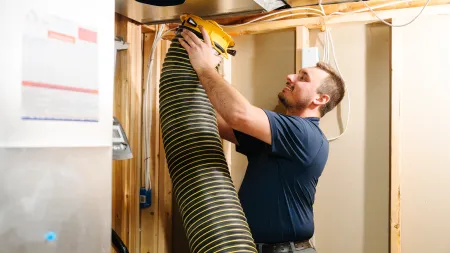 Technician installing flexible ductwork in a basement HVAC system with wood framing and insulation.