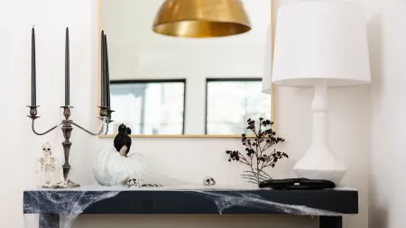 Halloween-themed mantel with black candles, skeleton, white pumpkin, crow, spider webs, and white lamp in modern room.