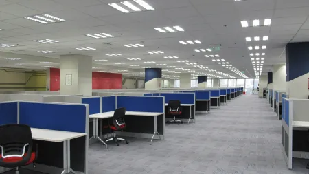 Modern office space with rows of blue cubicles and black chairs under bright fluorescent lighting.