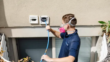 Technician wearing a respirator mask using a hose to clean exterior dryer vents on a house wall outdoors.