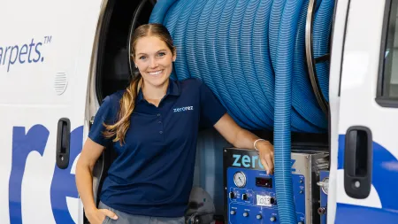 Smiling woman technician in blue uniform standing beside carpet cleaning equipment with coiled blue hoses in service van