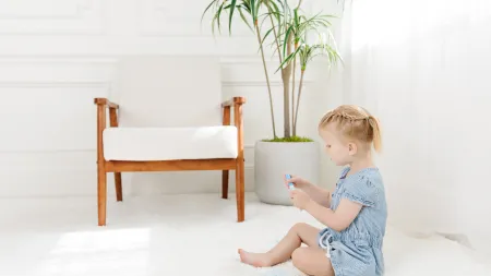 Young girl in casual dress playing on soft carpet beside a stylish chair and potted plant in bright room.