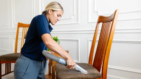 Woman vacuuming a brown upholstered wooden chair in a bright, white paneled room