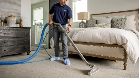 Professional cleaner uses carpet cleaning machine in a modern, well-lit bedroom with neutral decor.