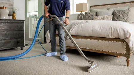 Professional technician cleaning carpet in a bright bedroom with modern decor and upholstered bed.