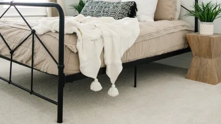 a bed with a white blanket with metal legs indenting in a beige carpet