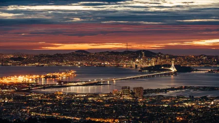 Nighttime cityscape of San Francisco with illuminated Bay Bridge and colorful sunset sky over the bay waters.