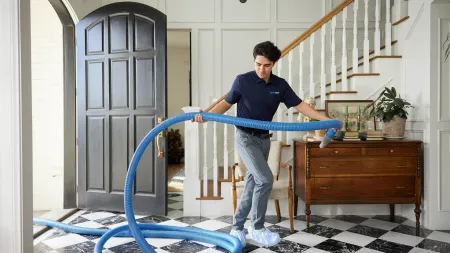 Professional technician in uniform inspecting a home interior with a large blue hose in a bright entryway.