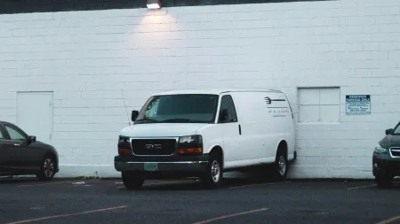 White GMC van parked in urban lot beside white and red brick buildings under overcast sky.