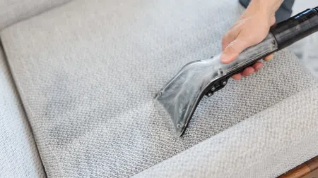 Hand using a vacuum cleaner attachment to clean a textured light gray couch.