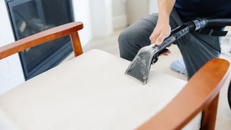 Professional cleaning of beige upholstered chair with vacuum upholstery tool and wooden armrests