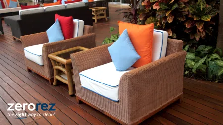 Outdoor wicker chairs with blue and orange pillows on wooden deck next to green plants and small table