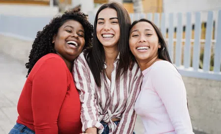 Three diverse young women smiling and hugging outdoors near a white fence, enjoying a sunny day together.