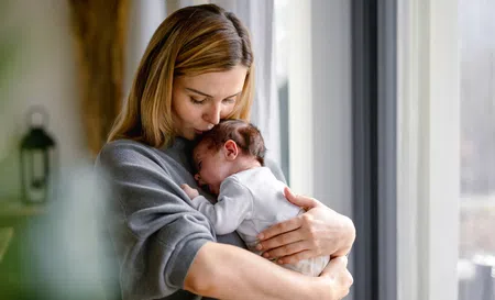 Mother tenderly holding and kissing newborn baby in cozy home by window with natural light.