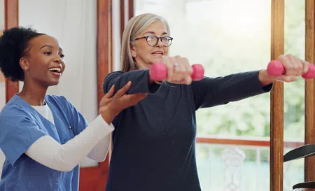Senior woman lifting pink dumbbells guided by a smiling female fitness trainer indoors near window.