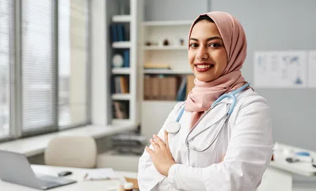 Young female doctor wearing a pink hijab and white coat with stethoscope smiling in a bright medical office.