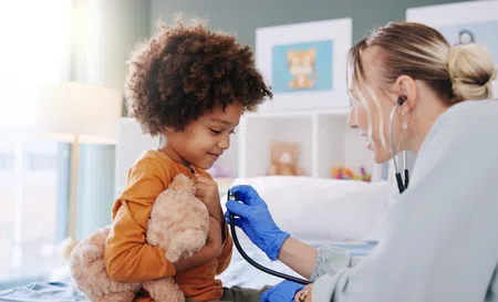 Child holding teddy bear while doctor listens to chest with stethoscope in bright clinic room