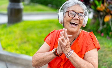 Joyful elderly woman in orange shirt wearing headphones and clapping hands outdoors in a garden.