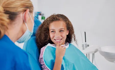 Smiling young girl sitting in dental chair wearing a blue bib while dentist in mask talks to her.