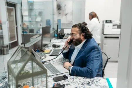 Man in blue suit talking on office phone while working on laptop in modern office environment.