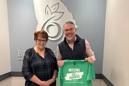 Two people standing indoors holding a green shirt with Missing 1000+ Pounds text, smiling in an office setting.