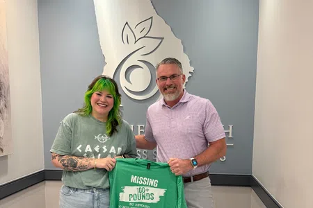 Two people in a modern office holding a green shirt that reads Missing 100+ Pounds support program by Body Experts.