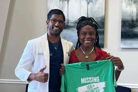 Smiling woman holding green shirt reading missing 100+ pounds with man in white coat giving thumbs up indoors.